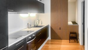 Eine moderne Küche mit dunklen Schränken, einer Edelstahl-Arbeitsplatte und einem eingebauten Backofen. Ein Holztrennwerk mit vertikalen Lamellen und eine kleine Sitzecke mit einer Bank und einem Tisch sind ebenfalls vorhanden.