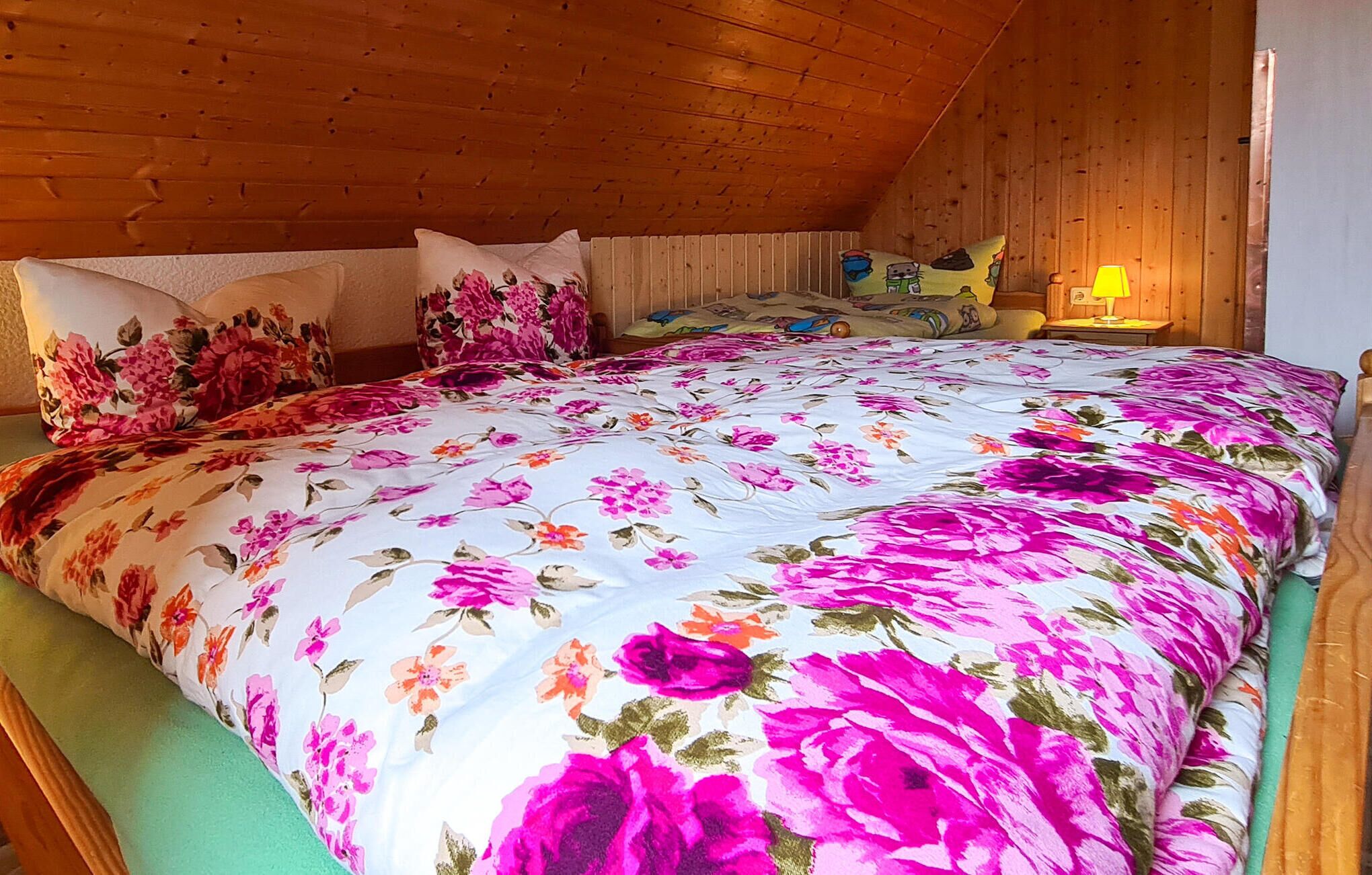 Un dormitorio con colcha de flores, paredes de madera, una mesita de noche con lámpara y una ventana con vistas al exterior.