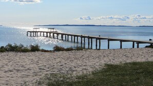 Una spiaggia con un molo di legno che si protende su acque calme, circondata da sabbia e vegetazione rada.