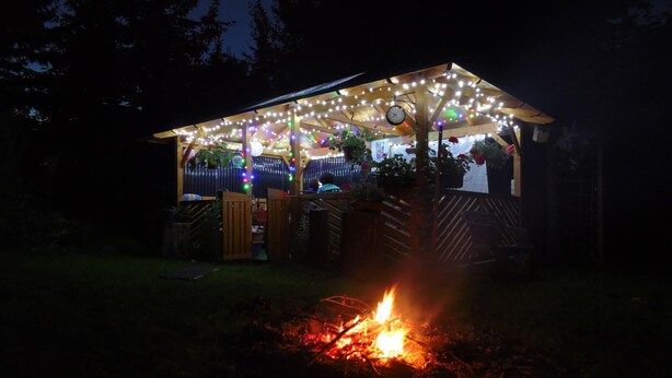 Une structure en bois décorée de guirlandes lumineuses et ornée d’une horloge, entourée de verdure et d’un feu de camp.