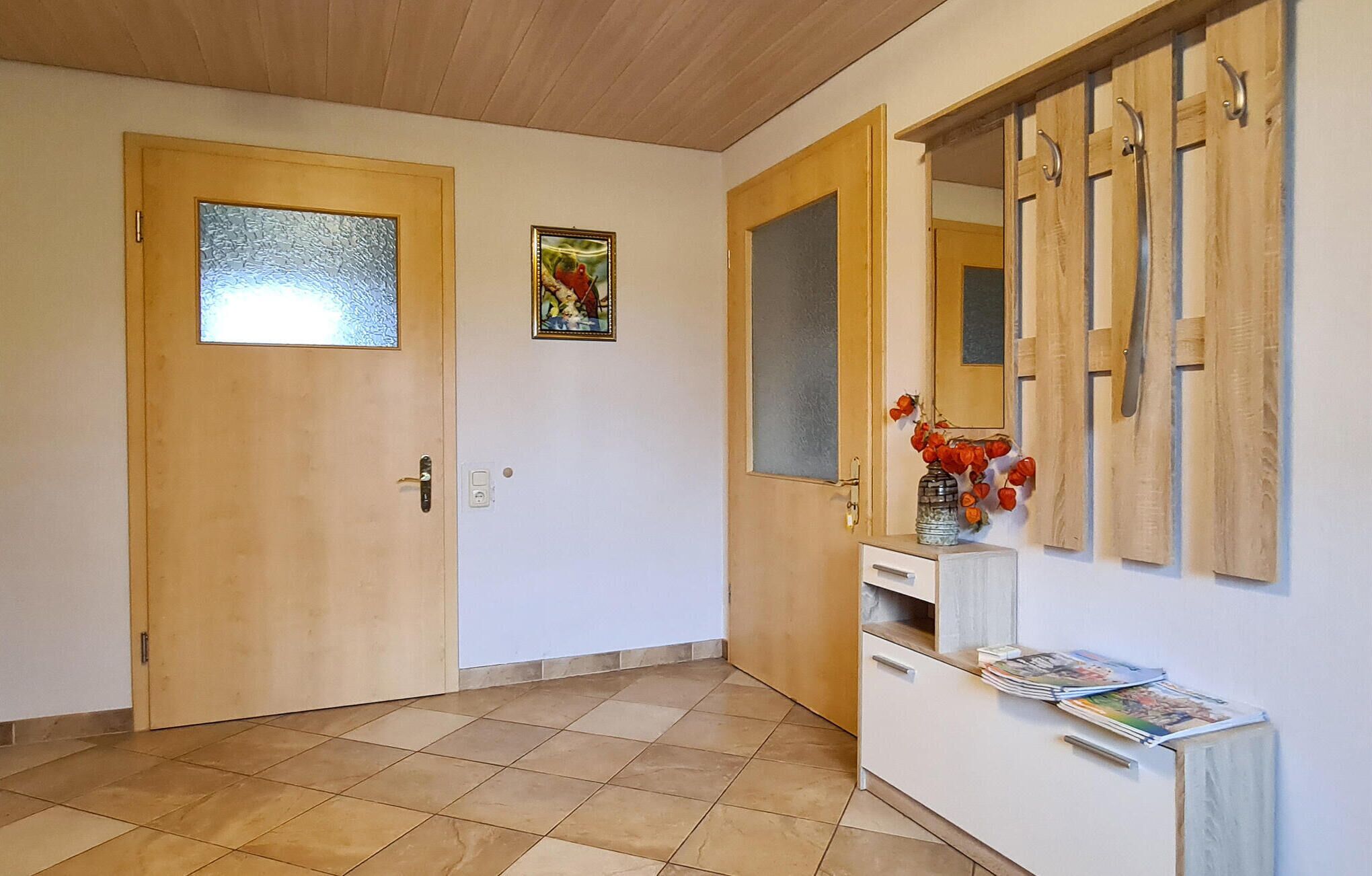 A hallway with two wooden doors, one with a frosted glass panel, a small wall-mounted shelf with a vase and magazines, and a tiled floor.