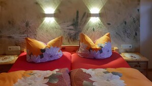 Une chambre avec deux lits, chacun agrémenté de draps à motifs floraux et de coussins décoratifs. On y trouve des lampes fixées au mur et une petite table avec un lampadaire.