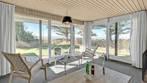 Amplia habitación con ventanas grandes que ofrecen vista a la playa y al océano. Hay una mesa de madera con una botella de Coca-Cola, un cuenco de palomitas de maíz y una vela.