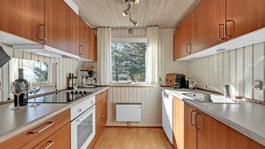Cocina compacta con armarios de madera, horno integrado, cafetera y ventana con vista a árboles.