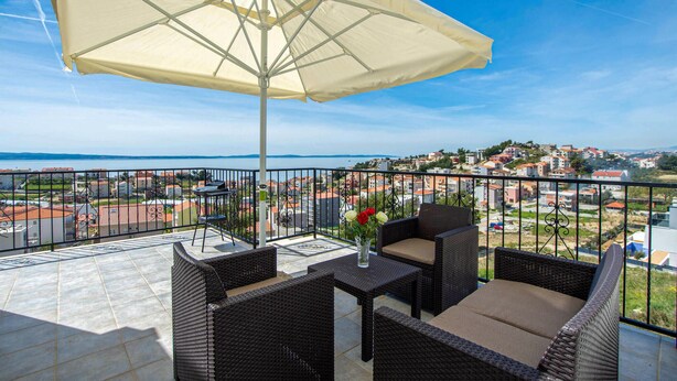 A balcony with wicker furniture, a table, and an umbrella, overlooking a coastal town. - Nice apartment in Podstrana with WiFi (Podstrana)