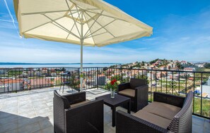Varanda com mobiliário de vime, uma mesa e um guarda-sol, com vista para uma cidade costeira.