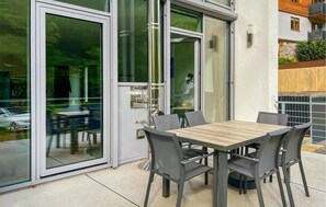 Eine Terrasse im Freien mit Tisch und Stühlen, eine Glastür und Blick auf ein parkendes Auto und Grünflächen.