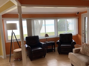 A living room with two leather armchairs, a small table, and a floor lamp. Large windows with blinds offer a view of the outdoors.