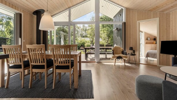 A spacious room with a high ceiling, large windows, a dining area with wooden furniture, and a cozy seating area with a fur throw. - 3 bedroom beautiful home in Glesborg (Glesborg)