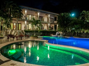 A swimming pool with a curved design, a lounge area with chairs, and a multi-story building with balconies and lighting. - Hotel Amira (Puerto Carrillo)