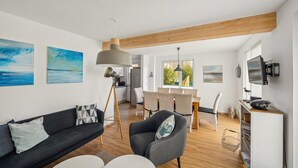 A modern living room with a dark sofa, a grey armchair, and a dining area with a table and chairs. There are paintings on the walls and a TV mounted on the wall. - Beautiful home in Süssau/Ostsee with sauna (Süssau/Ostsee)