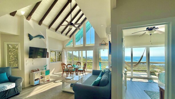 A spacious living room with a large window overlooking a scenic view, a ceiling fan, and a dining area with chairs and a table. - Luxury Ocean View Villa in West Bay Roatan. 3 Bedrooms with Pool & Beach Access (Roatan)