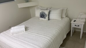 A neatly made bed with a white quilt, a white pillow with a black design, and a white nightstand with a remote control and a rolled-up towel. - Vamos a la playa, private pool/central AC (Vega Alta)