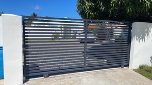 A modern metal gate with horizontal slats, a concrete wall, and a blue container to the left. - Vamos a la playa, private pool/central AC (Vega Alta)