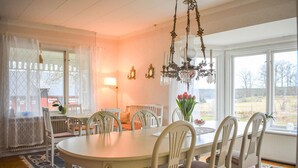 Een eetkamer met een grote ovale tafel, witte stoelen, een kroonluchter en uitzicht op het landschap door grote ramen.