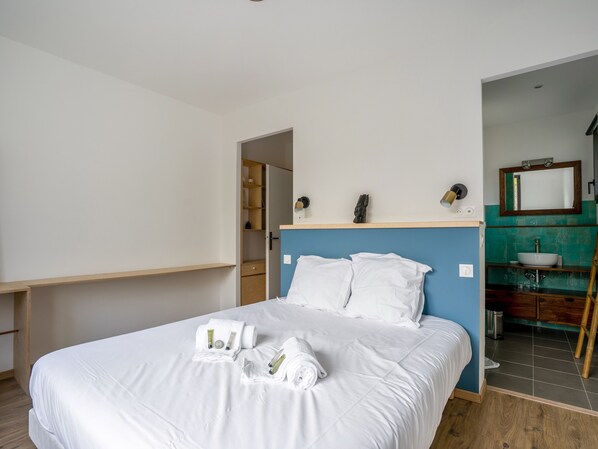 A hotel room with a bed, a wooden nightstand, a blue headboard, a bathroom with a sink and mirror, and a wooden floor.
