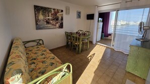 A room with a sofa, a dining table with chairs, a painting on the wall, and a view through a window. - 2 Room apartment with pool Richelieu beach Cap d'Agde (LE CAP D AGDE)