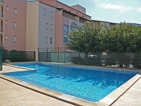 A rectangular swimming pool with a tiled deck, surrounded by a fence and a building with multiple windows and balconies.