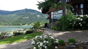 Un chalet au bord du lac, avec un sentier de pierres menant à une terrasse en bois, entouré d’une végétation luxuriante et dominé par une chaîne de montagnes.
