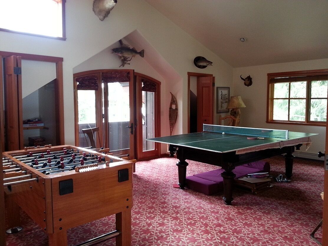 A room with a foosball table, ping pong table, and a large window with a view of trees.