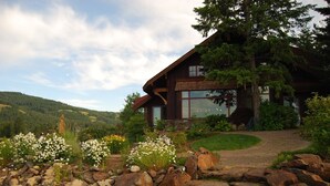 Un chalet rustique, doté de fondations en pierre, entouré d’un jardin où poussent des fleurs blanches et dont un sentier de pierres mène à l’entrée.