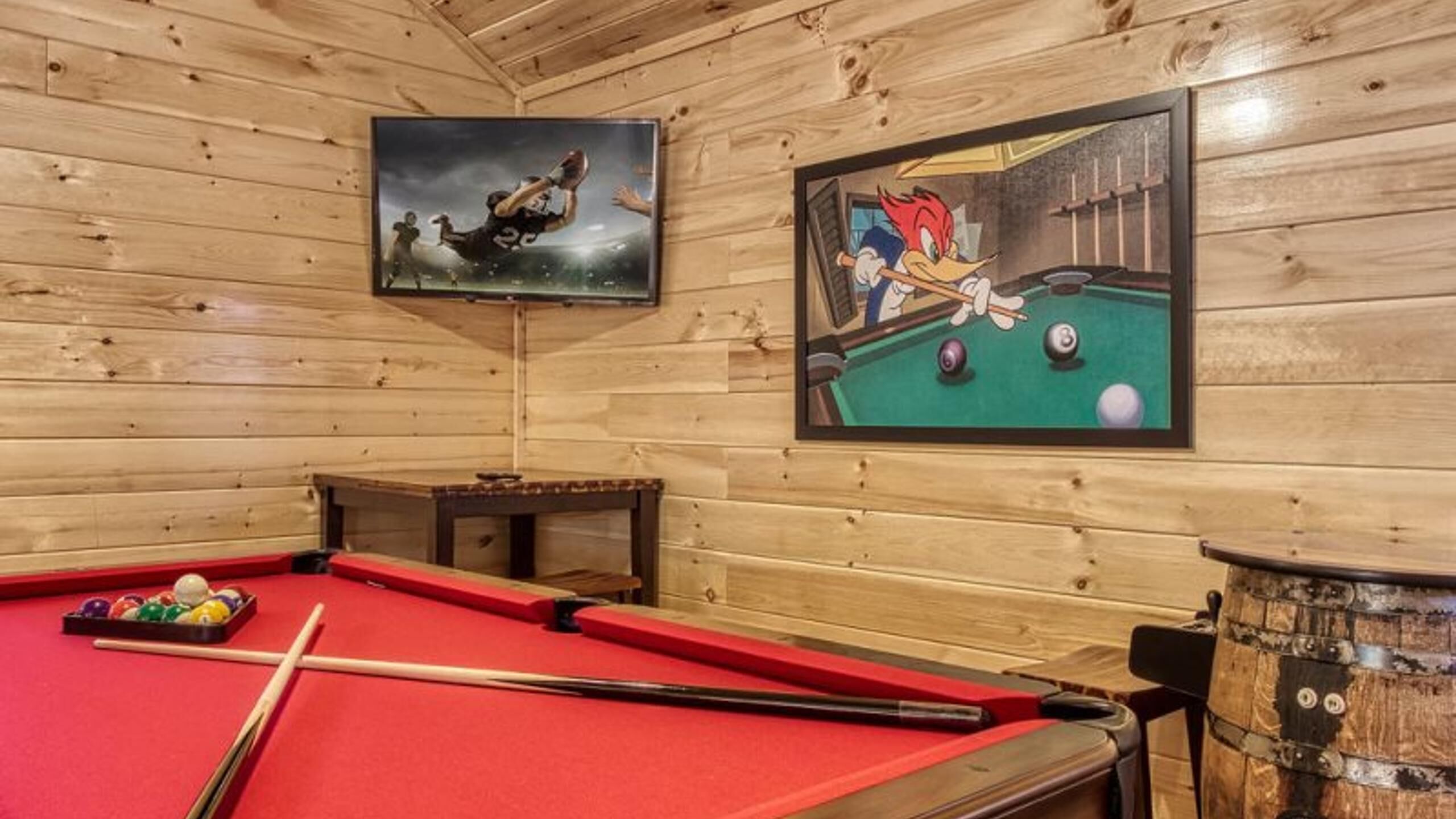 A pool table with a red felt surface, a pool cue, and a set of balls. There are two framed pictures on the wall, one depicting a football player and the other a cartoon character playing pool.