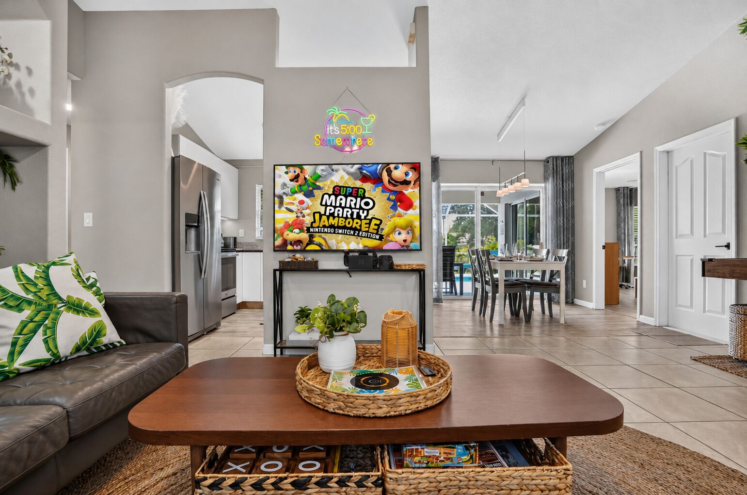 A modern living room with a flat-screen TV, a wooden coffee table, and a dining area in the background.