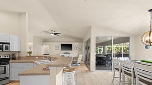 Una cocina moderna con una isla central, una zona de comedor con sillas blancas y vistas a un patio exterior a través de puertas de cristal.