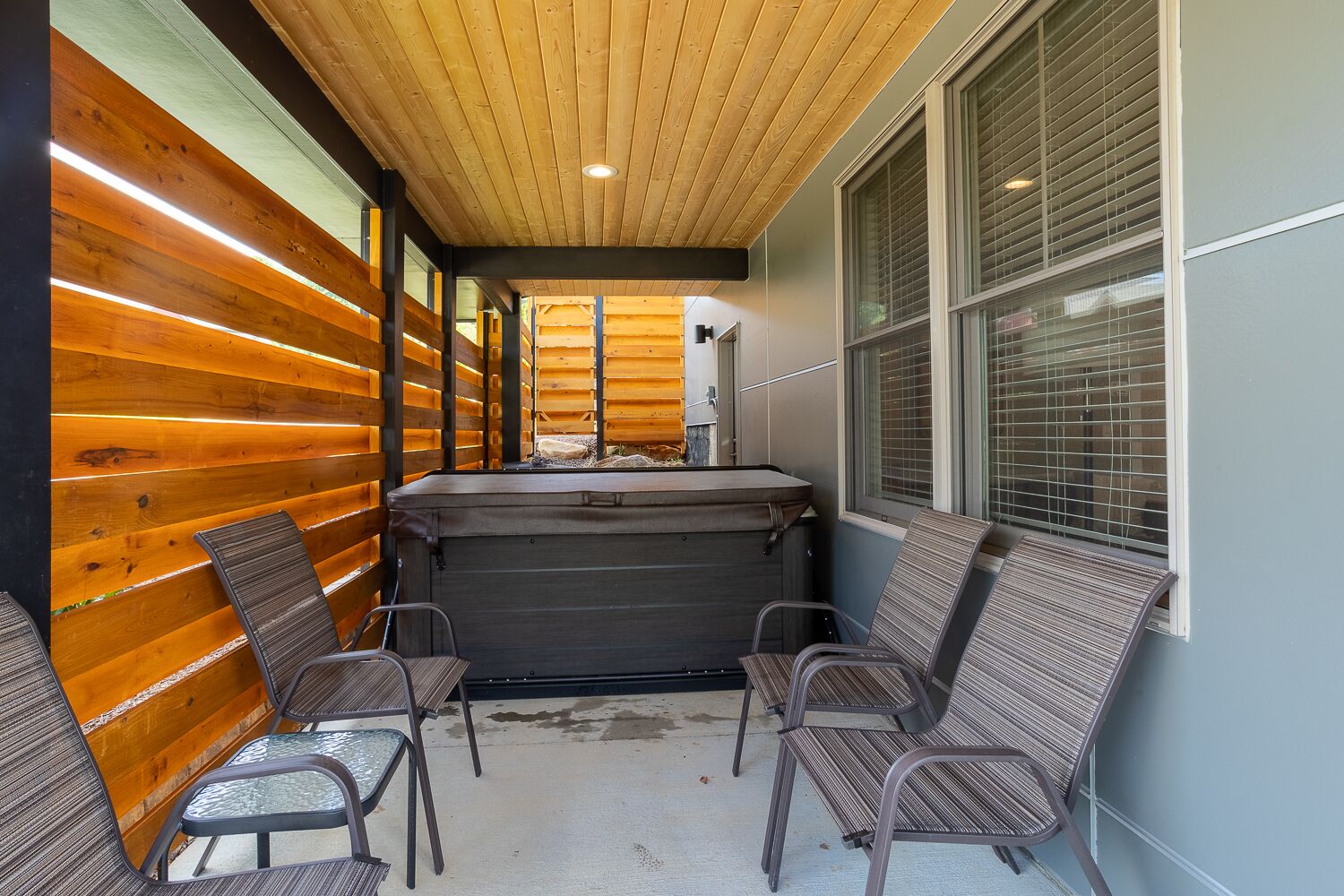 A covered outdoor seating area with a hot tub, wooden walls, and metal chairs.