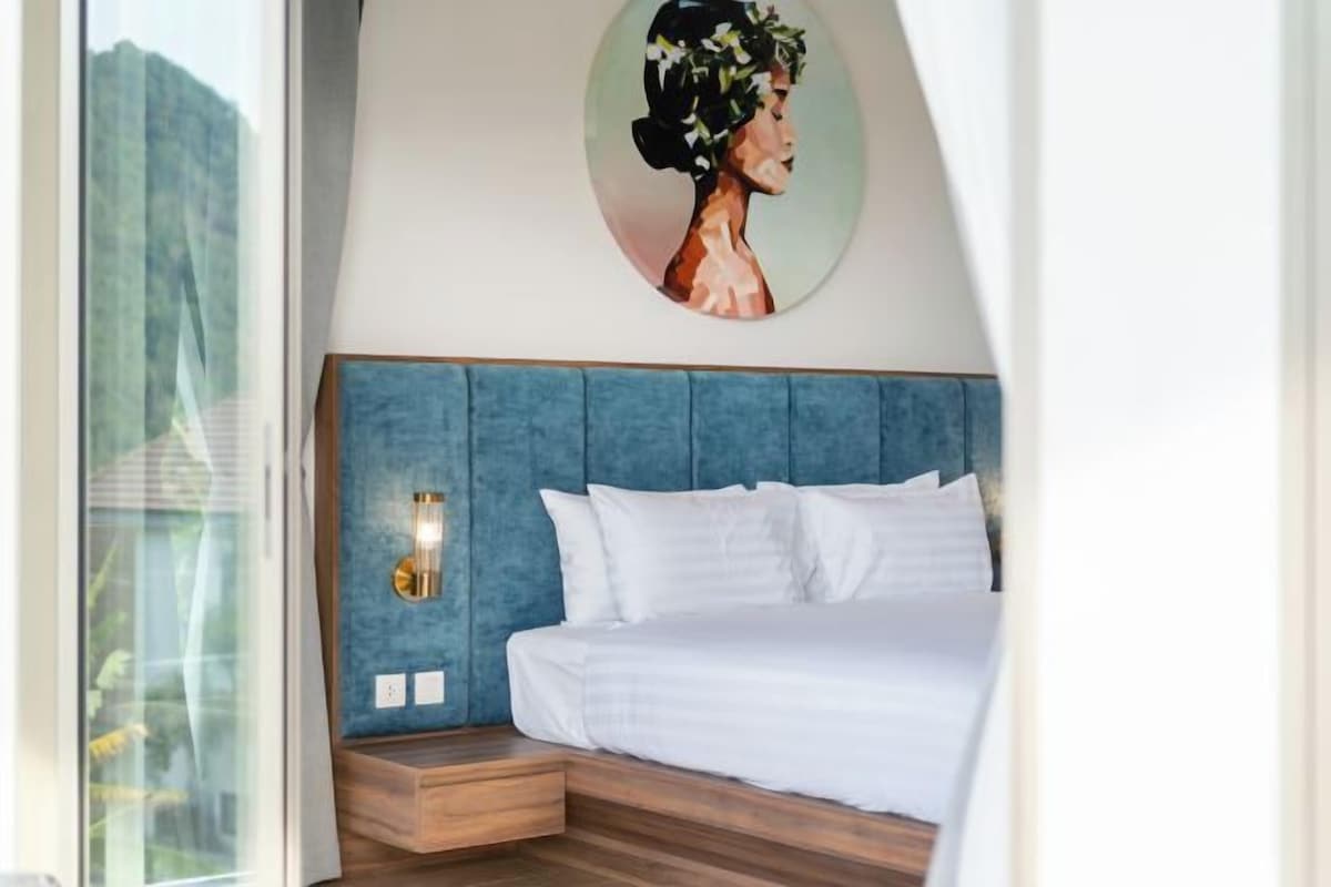 A modern bedroom with a blue headboard, a bed with white linens, a wall-mounted light, and a framed artwork above the bed.
