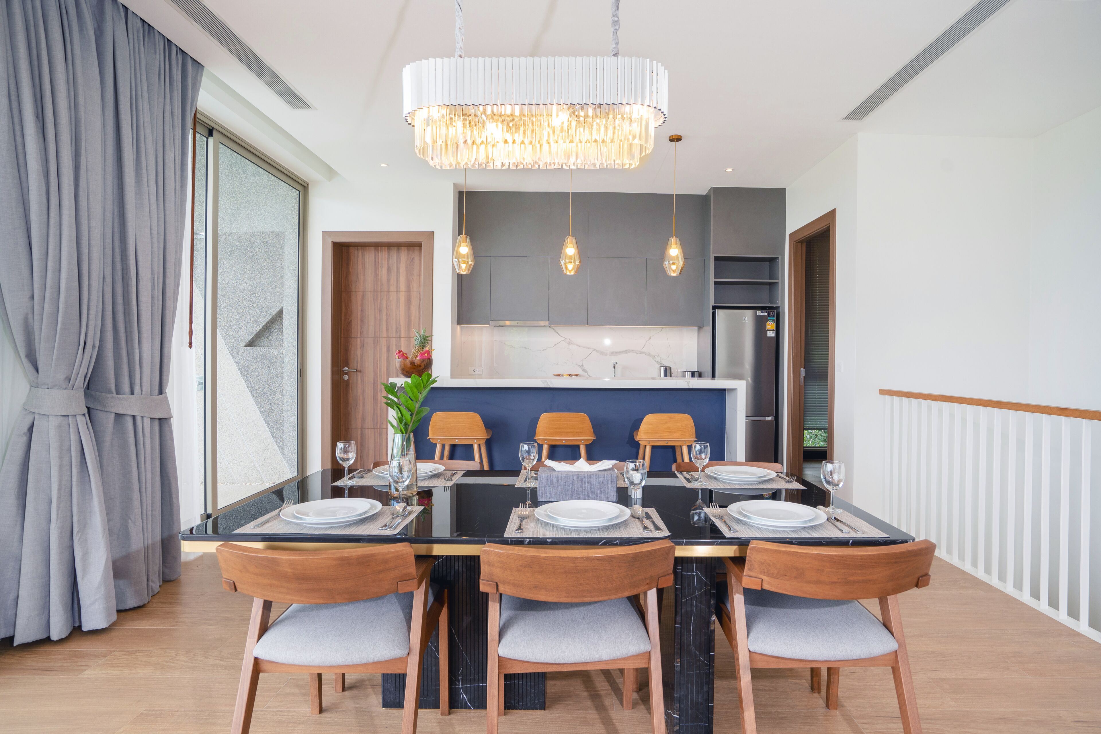 A modern dining area with a large table set for six, wooden chairs, a chandelier, and a kitchen in the background.