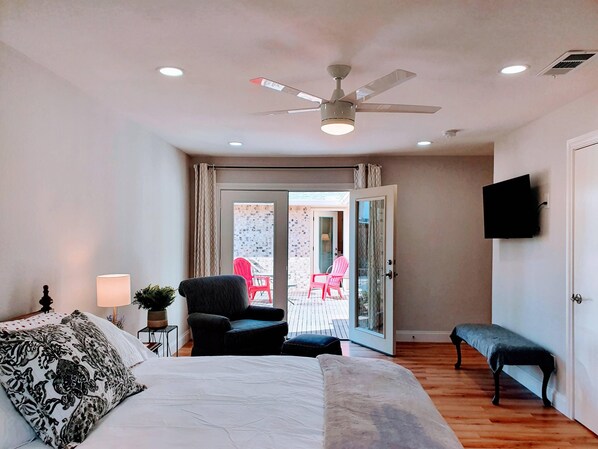 A bedroom with a bed, a ceiling fan, a bench, a TV, and a sliding glass door leading to an outdoor area with chairs.