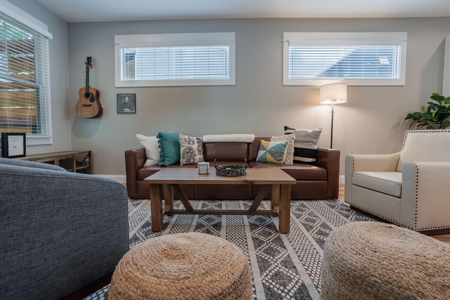 Un salón acogedor con un sofá de cuero marrón, un sillón gris y una mesa de centro de madera. Se ven dos ventanas con persianas, una guitarra apoyada en la pared y una lámpara a la derecha.