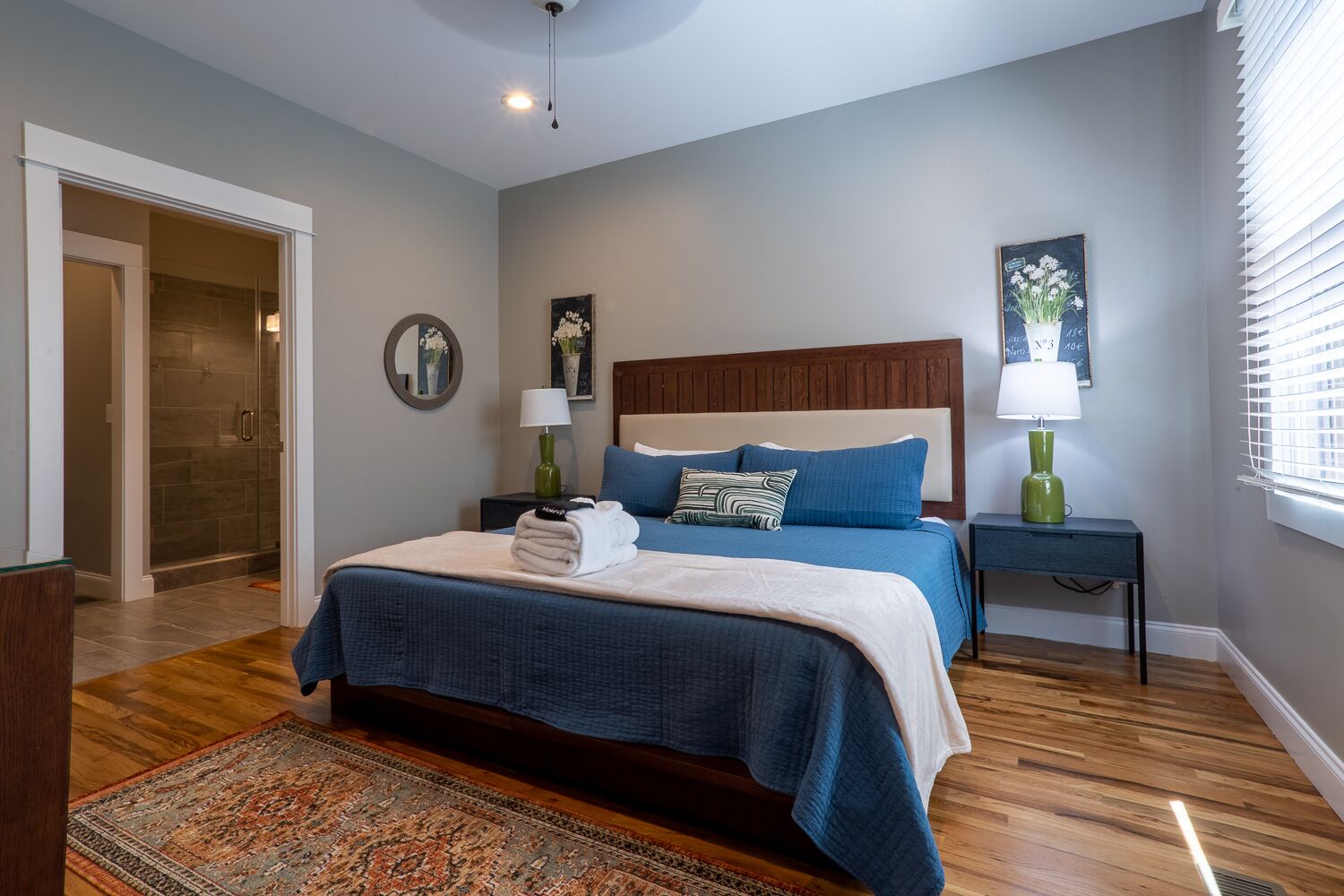 A bedroom with a large bed, two bedside tables, a nightstand, a mirror, and a window with blinds.