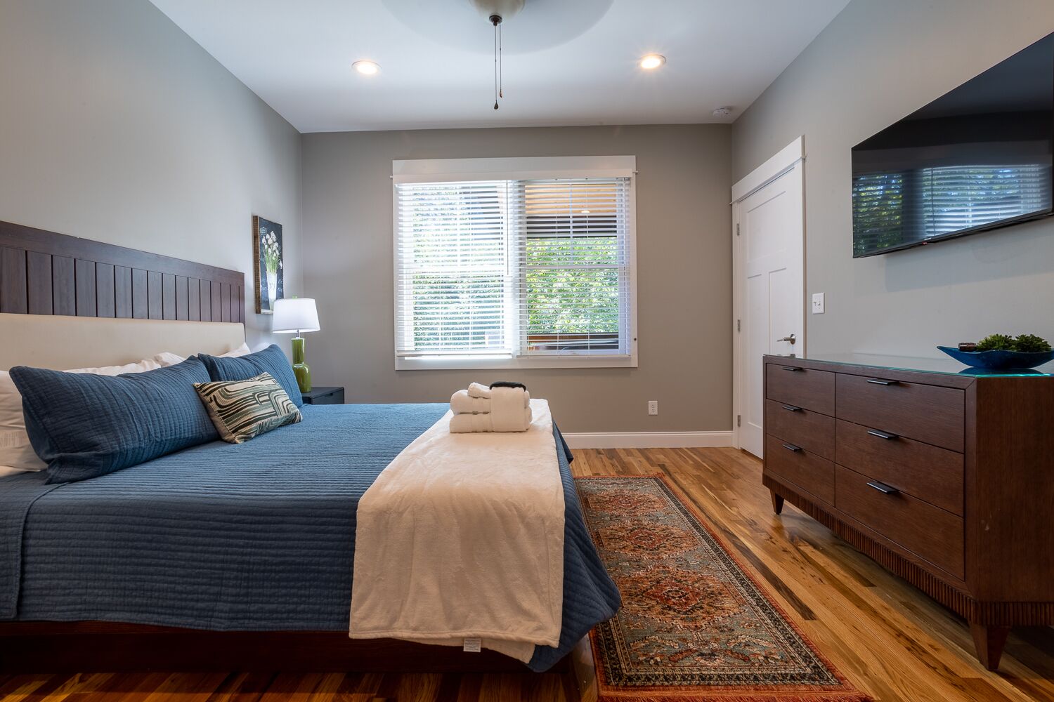 A bedroom with a bed, bedside table, lamp, window with blinds, dresser, and a television on the wall.