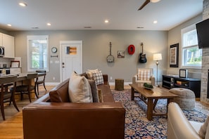 A modern living room with a brown leather sofa, a wooden coffee table, and a patterned rug. There is a dining area with a kitchen visible in the background.