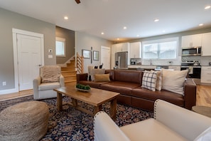 A modern living room with a brown leather sofa, white armchairs, and a wooden coffee table. A staircase leads to the upper floor.