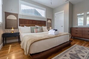 A bedroom with a large bed, a nightstand with a lamp, a dresser, and a window with blinds.