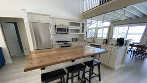 Eine moderne Küche mit einer großen Holzinsel, schwarzen Barhocker, Edelstahlgeräten und Blick ins Freie durch große Fenster.