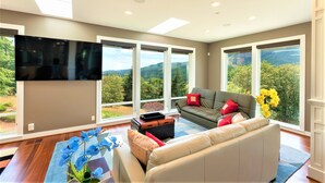 A spacious living room with a large flat-screen TV, multiple seating options, and large windows offering a scenic view. - New Listing - Breathtaking vacation/work remotely in wine country! (Forest Grove)
