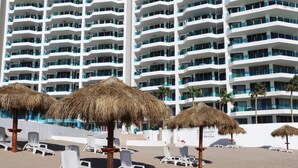 Una playa con sillas de descanso blancas y sombrillas de paja, un hotel de gran altura con balcones al fondo.