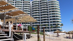 Un hotel frente a la playa con varios balcones, una zona de bar y una playa de arena con sombrillas.