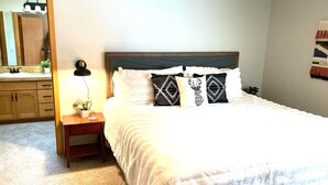 Ein ordentlich bezogenes Bett mit weißer Bettwäsche, einem Holz-Kopfteil und dekorativen Kissen. Ein Nachttisch mit Lampe und einer kleinen Pflanze steht neben dem Bett. Im Hintergrund sind ein Spiegel und ein Holzschrank zu sehen.