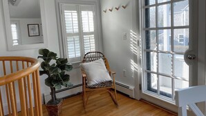 Una habitación con piso de madera, una silla blanca con cojín, una cuna de madera, una planta en maceta y un amplio ventanal con persianas.