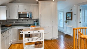 Cocina con armarios blancos, microondas, mesa de madera con un cuenco de fruta y una lámpara colgante.