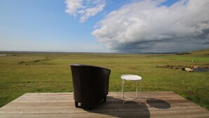 A wooden deck with a black chair and a small white table, overlooking a grassy field and a cloudy sky.