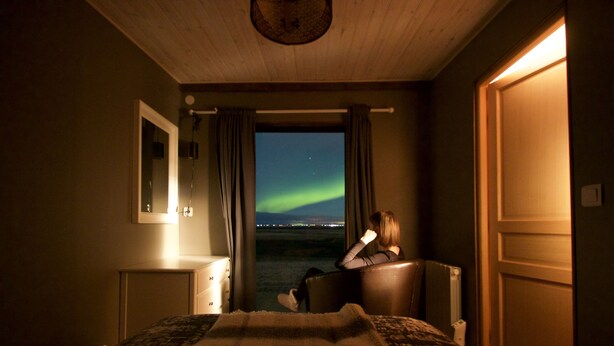 A bedroom with a bed, a nightstand, a mirror, a window with curtains, and a view of the night sky.