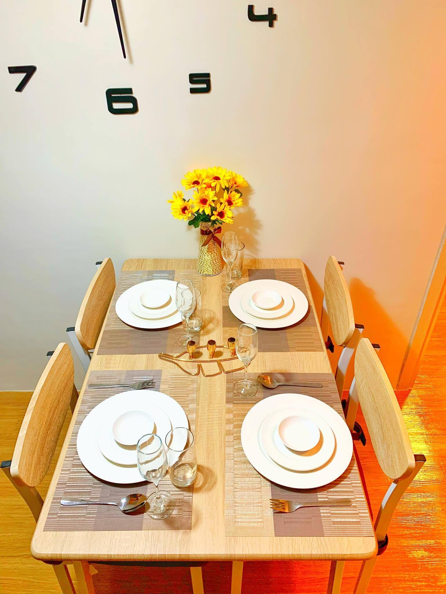 A dining table set for four with white plates, glasses, and cutlery, a vase with yellow flowers, and a clock on the wall.