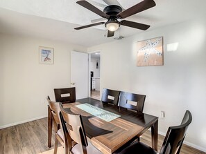 Une salle à manger avec une table en bois, quatre chaises, un ventilateur de plafond et des œuvres d’art accrochées aux murs.