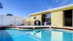 A swimming pool with a tiled deck, a covered grill, and a patio area with chairs and a table.